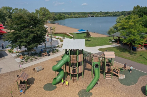 high angle over a children's playground with a slide complex sitting on brown mulched ground. There's a corner of a lake in the background